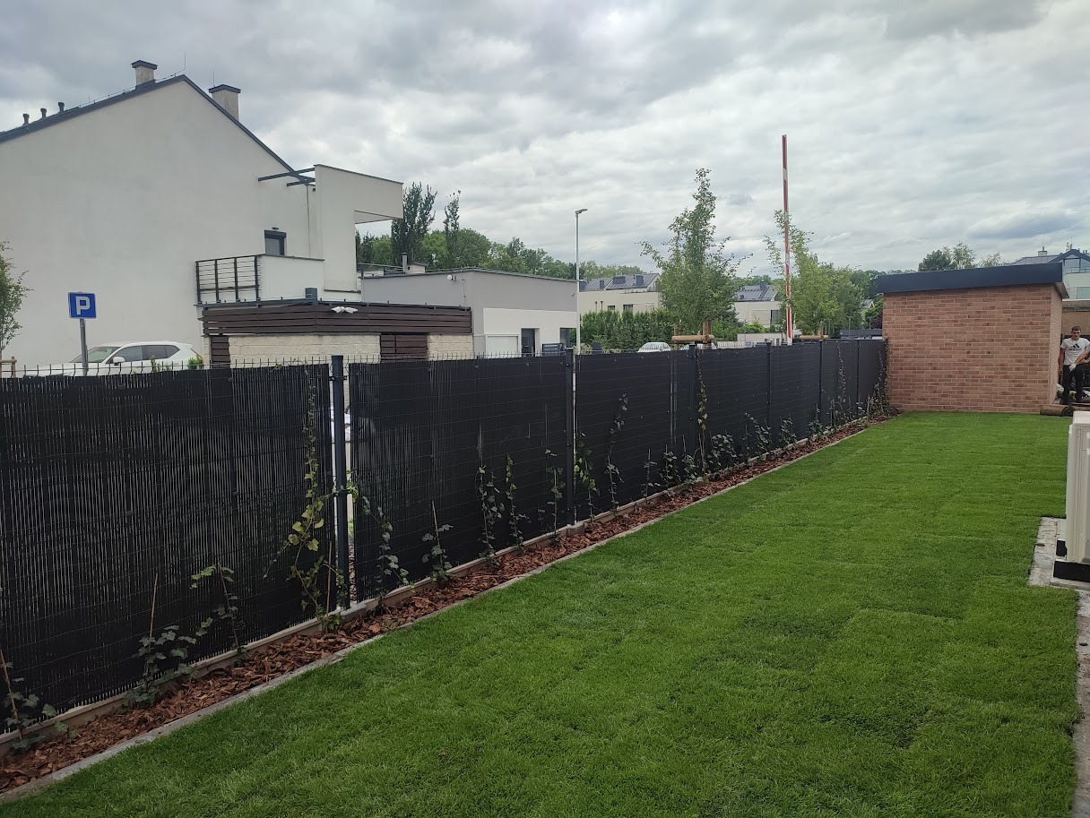 Nowo założony trawnik z rolki przylegający do czarnego ogrodzenia z pnącymi się roślinami, obrzeże wyłożone korą, w tle nowoczesny budynek mieszkalny.