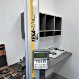 Wysokościomierz TESA Hite 700 na granitowym stole warsztatowym, obok drewniane pudełko z narzędziami pomiarowymi, w tle półka z przegródkami.