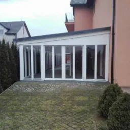 Zadaszony taras z białymi ramami okiennymi, przylegający do budynku z balkonem, wyłożony ażurowymi płytami z zielenią, otoczony tujami.