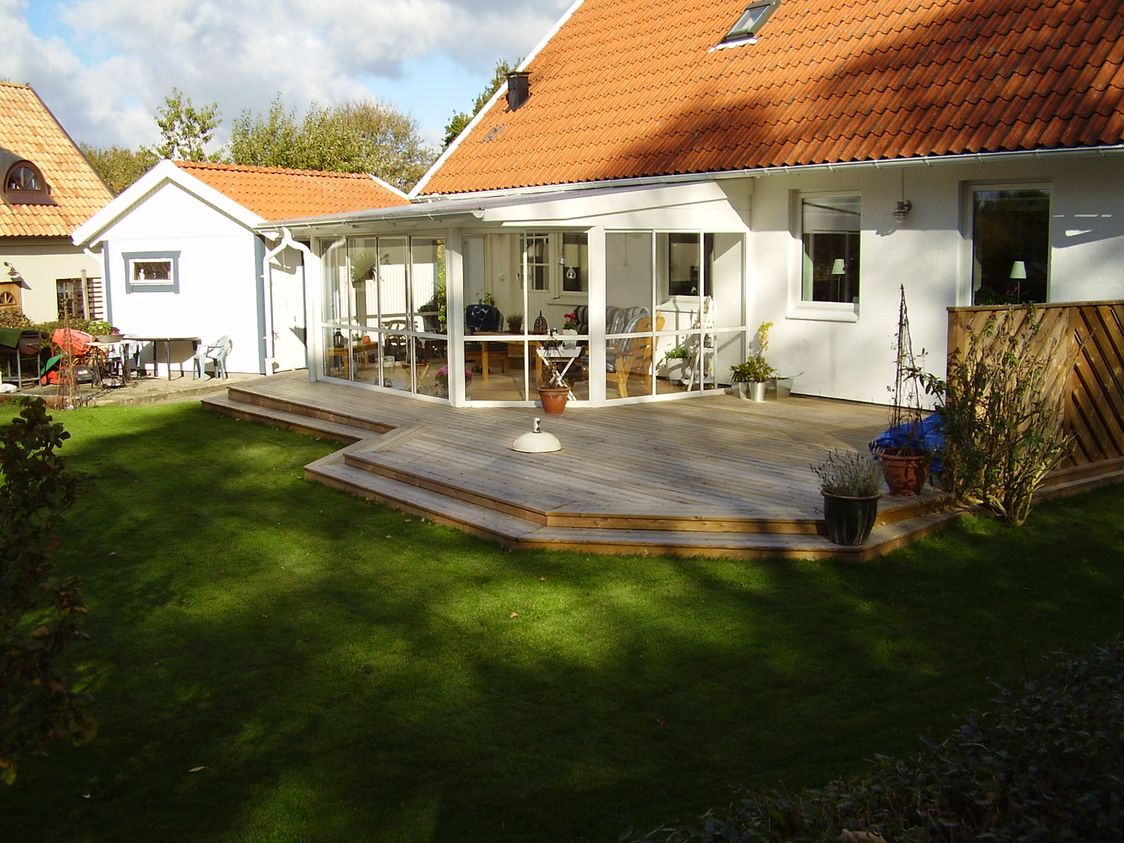 Jasny, drewniany taras z przeszkloną zabudową przylegający do białego domu z czerwoną dachówką. Widok na zadbany trawnik i część ogrodu w słoneczny dzień.