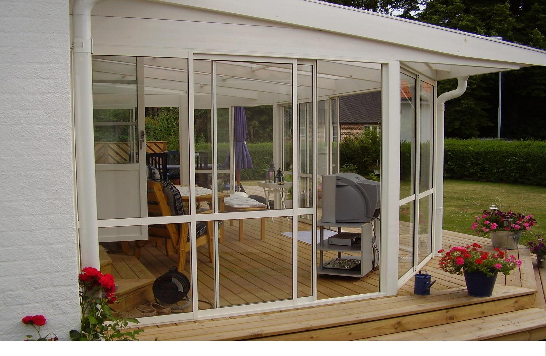 Biała zabudowa balkonu z przesuwnymi szklanymi drzwiami, drewnianym podestem i kwiatami w donicach. Wewnątrz widoczne meble i stary telewizor.