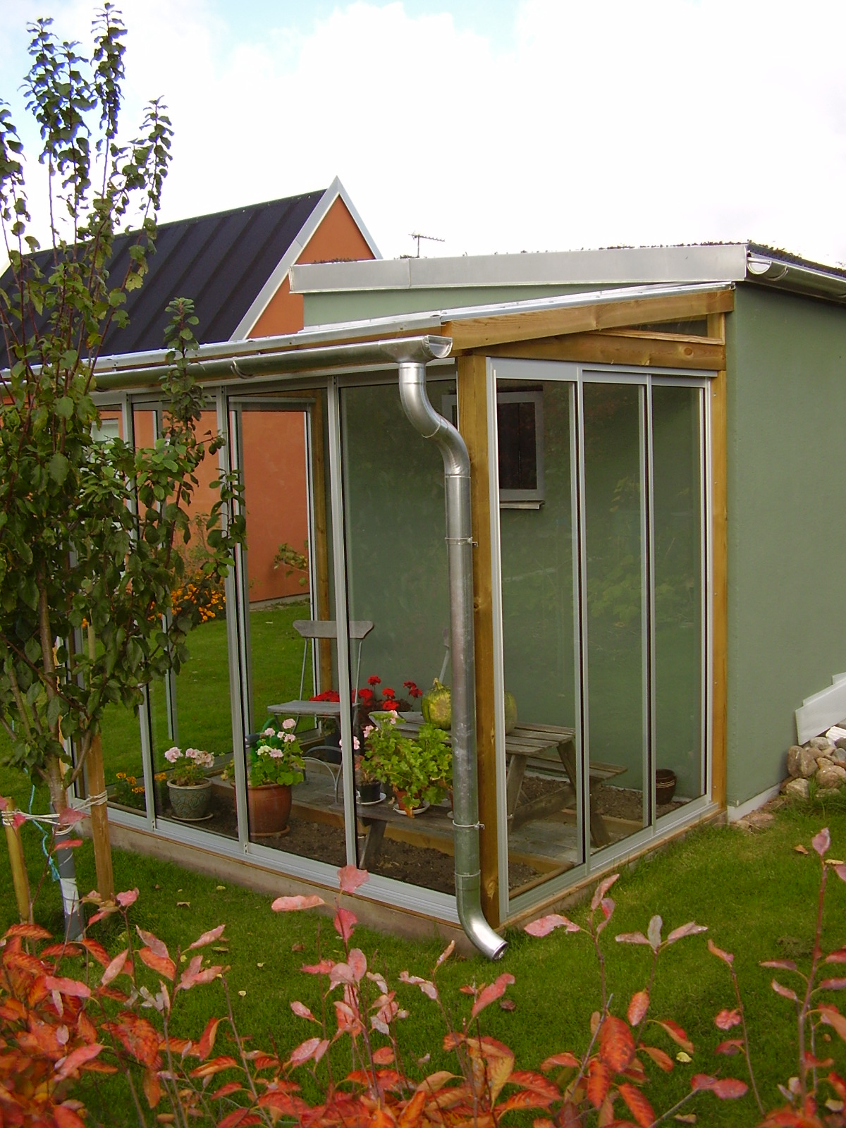 Szklana konstrukcja przylegająca do budynku z zielonymi ścianami. Wewnątrz rośliny w donicach, krzesła i ławka. Na zewnątrz trawnik i roślinność.