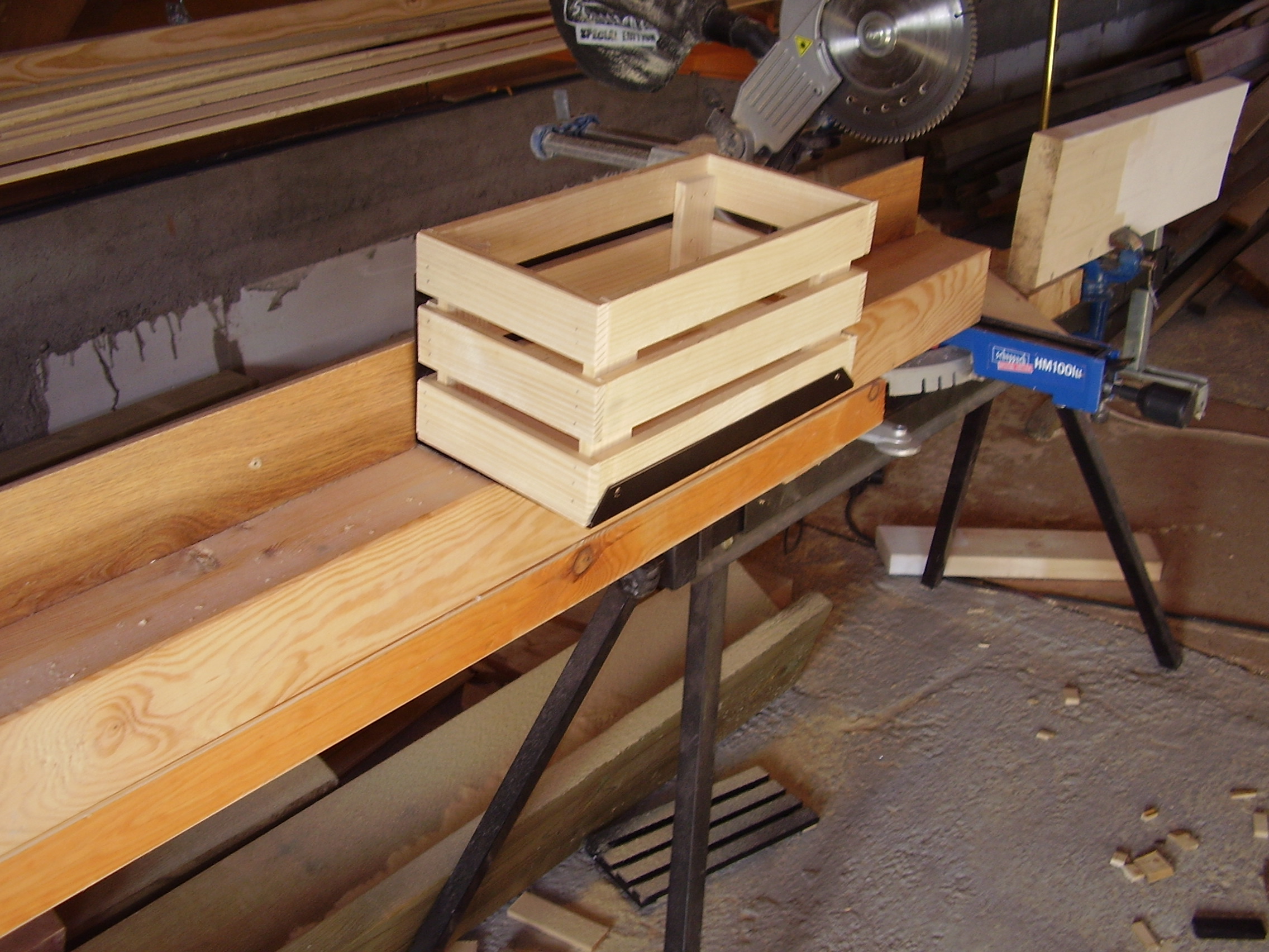 Drewniana skrzynka na stole warsztatowym, obok piła tarczowa i resztki drewna. Widoczne elementy konstrukcyjne i narzędzia stolarskie.