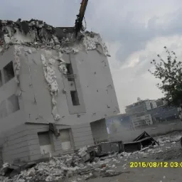 Demolka budynku w toku: fragment maszyny budowlanej kruszy górną część konstrukcji, gruz leży na ziemi. W tle zabudowa miejska i pochmurne niebo.