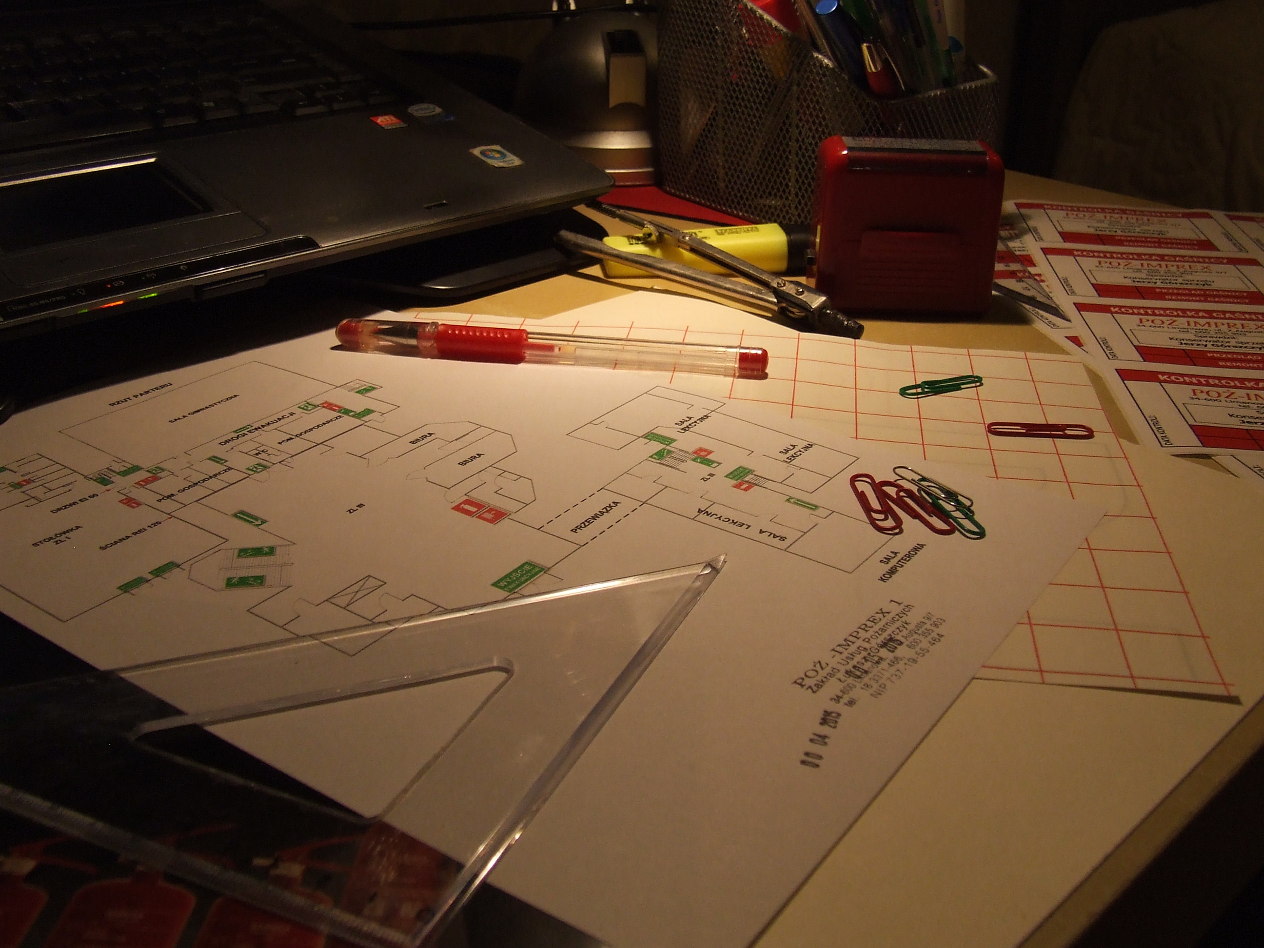 Plan ewakuacji budynku z zaznaczonymi drogami i wyjściami, leżący na biurku obok przyborów kreślarskich i pieczątek kontroli gaśniczej.