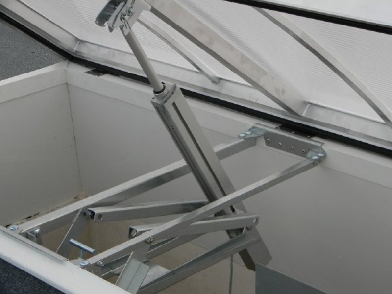 Otwarty świetlik dachowy z mechanizmem podnoszenia w Limanowej. Widoczne elementy konstrukcyjne i siłownik. Białe ściany i przezroczysty dach.