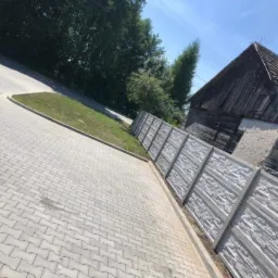 Nowoczesny betonowy płot panelowy z fakturą kamienia, oddzielający posesję od chodnika z kostki brukowej, z widocznym starym budynkiem w tle.