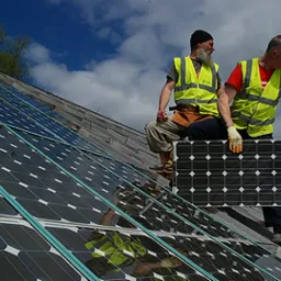 Dwóch monterów w kamizelkach odblaskowych instaluje panele słoneczne na dachu budynku w słoneczny dzień. Jeden z nich trzyma panel, a drugi siedzi na dachu.