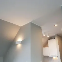 Wnętrze poddasza ze skośnym sufitem, oświetlone kinkietem ściennym i punktowymi lampami sufitowymi, widoczny fragment kuchni z białymi szafkami.