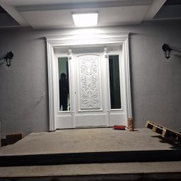 SpecTech Serwis - Nowo zamontowane, białe drzwi wejściowe z ozdobnymi elementami, widoczne na tle szarej ściany budynku i betonowych schodów, z paletą i narzędziami obok.