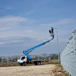 SpecTech Serwis - Pracownik na podnośniku montuje oświetlenie na słupie wzdłuż metalowego ogrodzenia. W tle krajobraz wiejski pod błękitnym niebem.