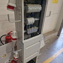 SpecTech Serwis - Otwarta, szara skrzynka elektryczna z widocznymi bezpiecznikami, przewodami i gniazdami przemysłowymi. Obok gaśnica i żółta kartka z napisem 'BRAMA 7'.