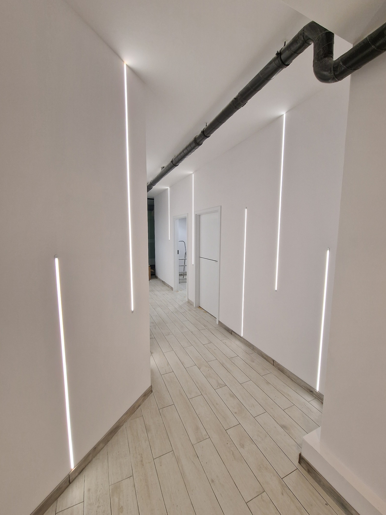 Nowoczesny korytarz z pionowymi liniami oświetlenia LED w ścianach, widoczne rury instalacyjne pod sufitem oraz podłoga z jasnych płytek imitujących drewno.