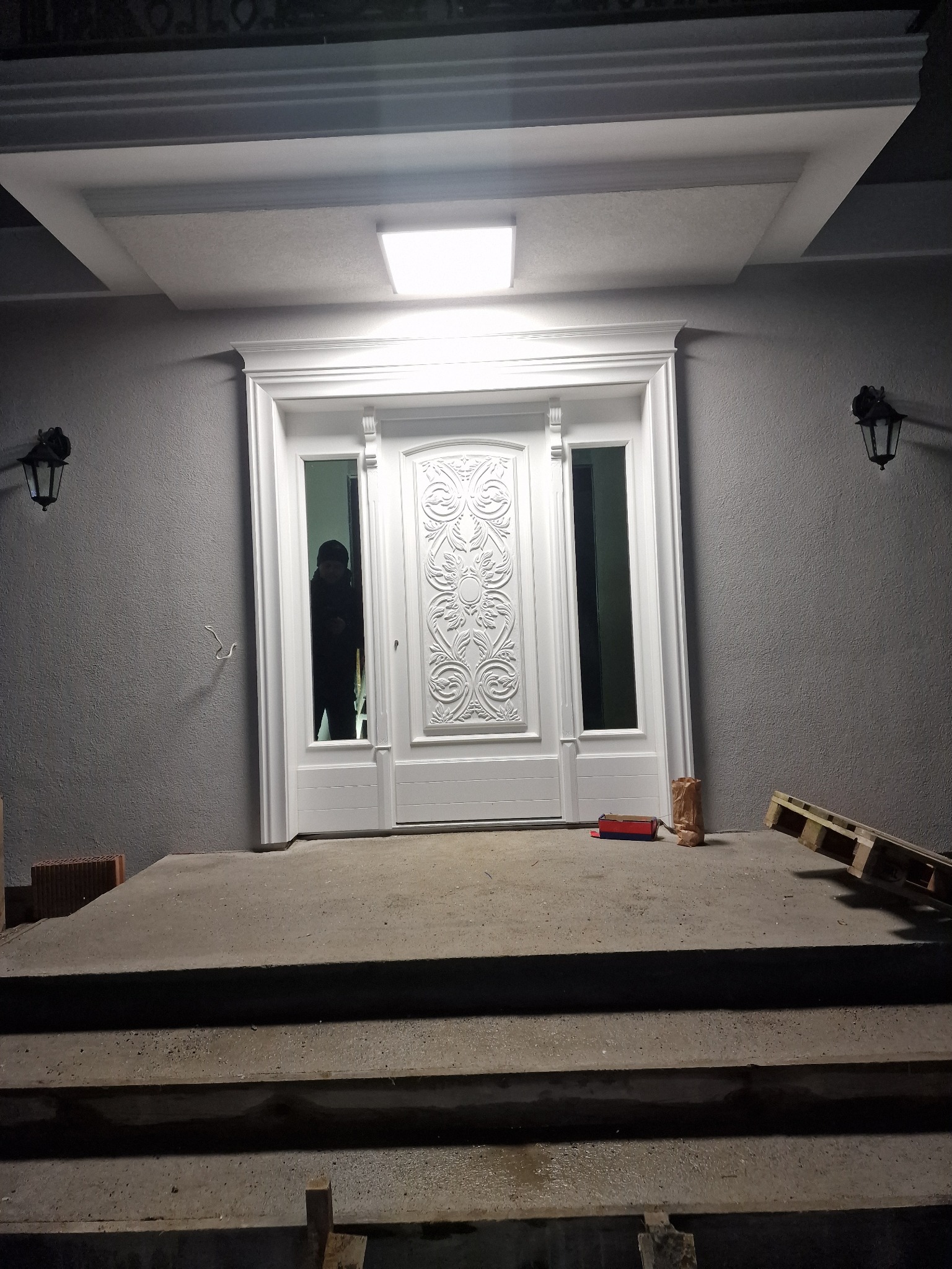 Nowo zamontowane, białe drzwi wejściowe z ozdobnymi elementami, widoczne na tle szarej ściany budynku i betonowych schodów, z paletą i narzędziami obok.