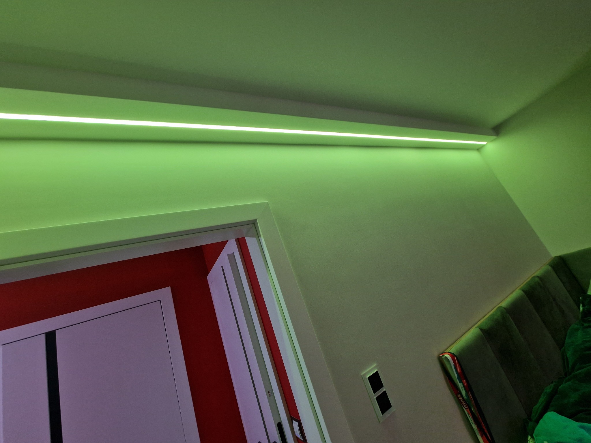Wnętrze pokoju z oświetleniem LED w postaci cienkiej listwy zamontowanej pod sufitem, dającej zielone światło, widoczny fragment ściany, drzwi i włącznik światła.