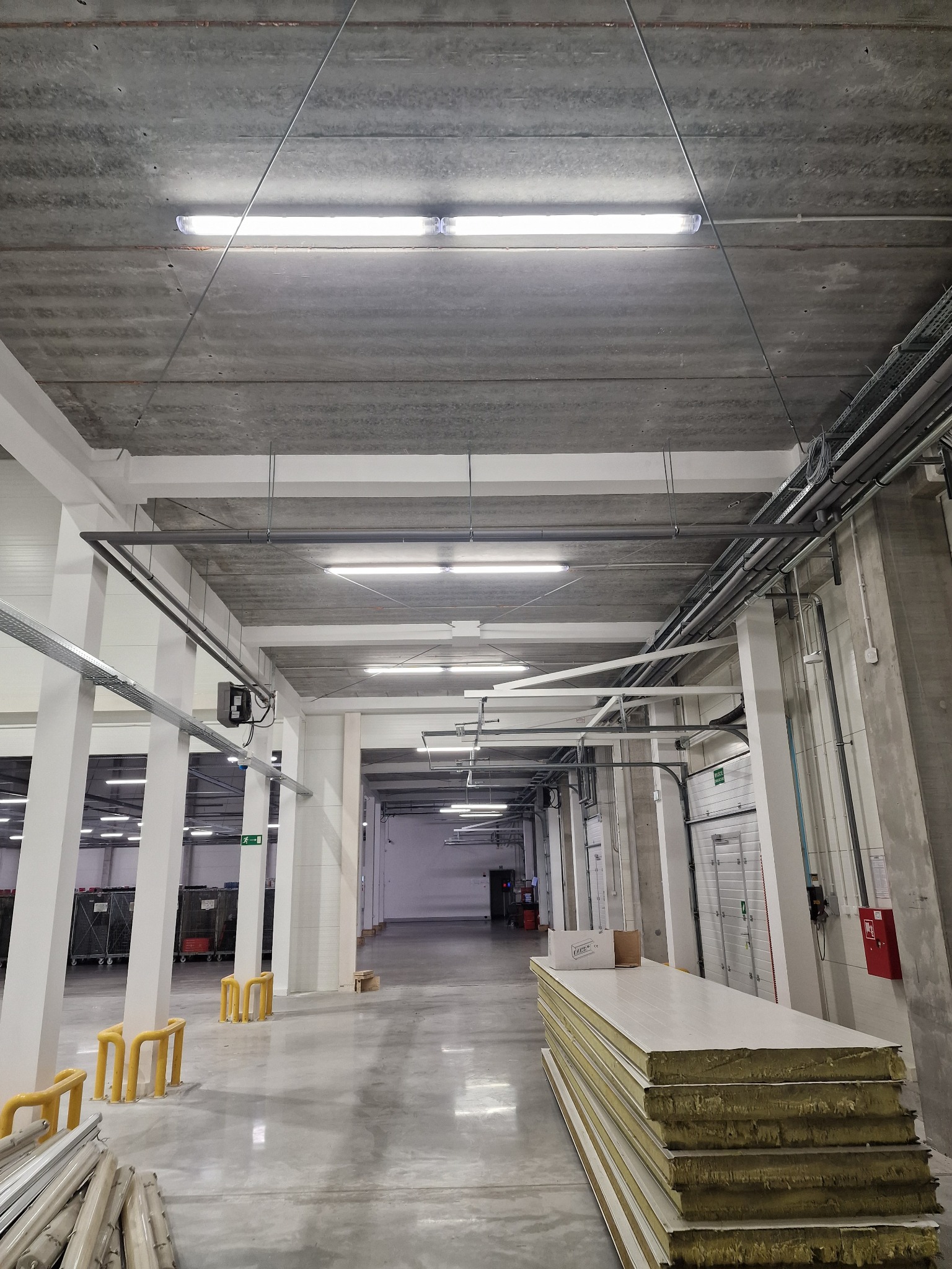 Wnętrze hali magazynowej z widocznymi rzędami świetlówek pod betonowym sufitem, instalacjami elektrycznymi na ścianach i stropie, oraz stosem płyt warstwowych na pierwszym planie.