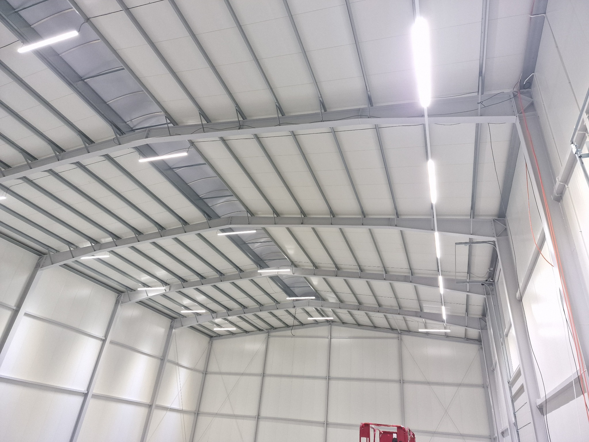 Wnętrze hali magazynowej z widoczną konstrukcją stalową dachu i zamontowanym oświetleniem LED w postaci podłużnych lamp.