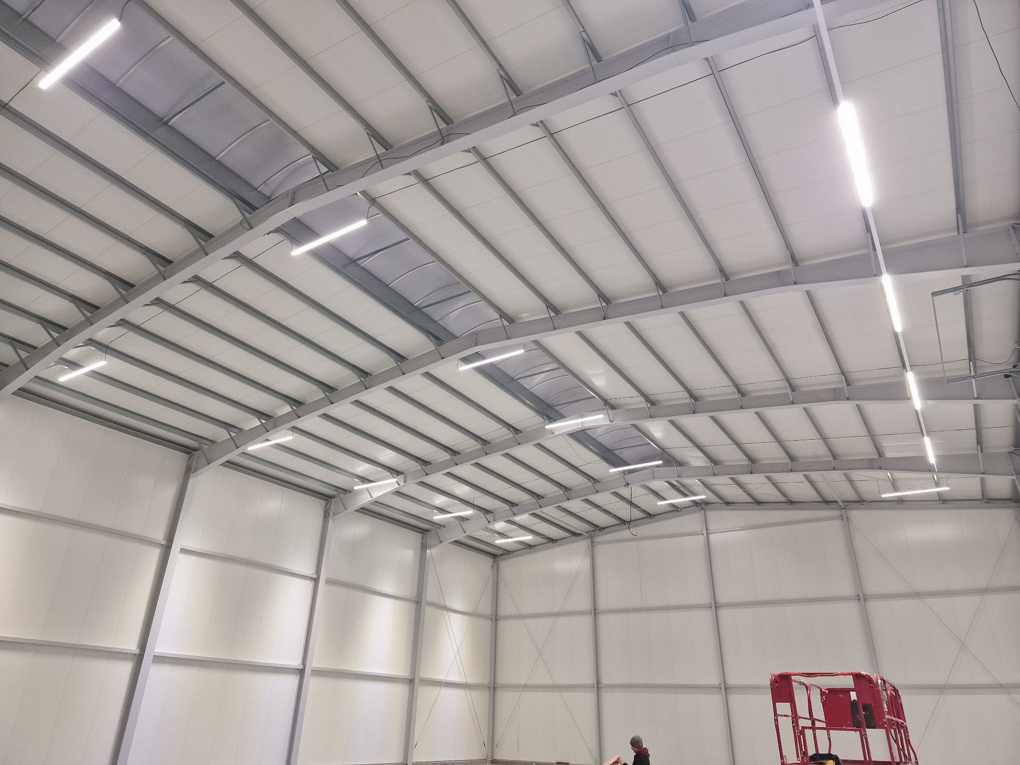 Wnętrze hali przemysłowej z zamontowanymi liniowymi lampami LED pod sufitem, widoczny podnośnik koszowy i pracownik w oddali.