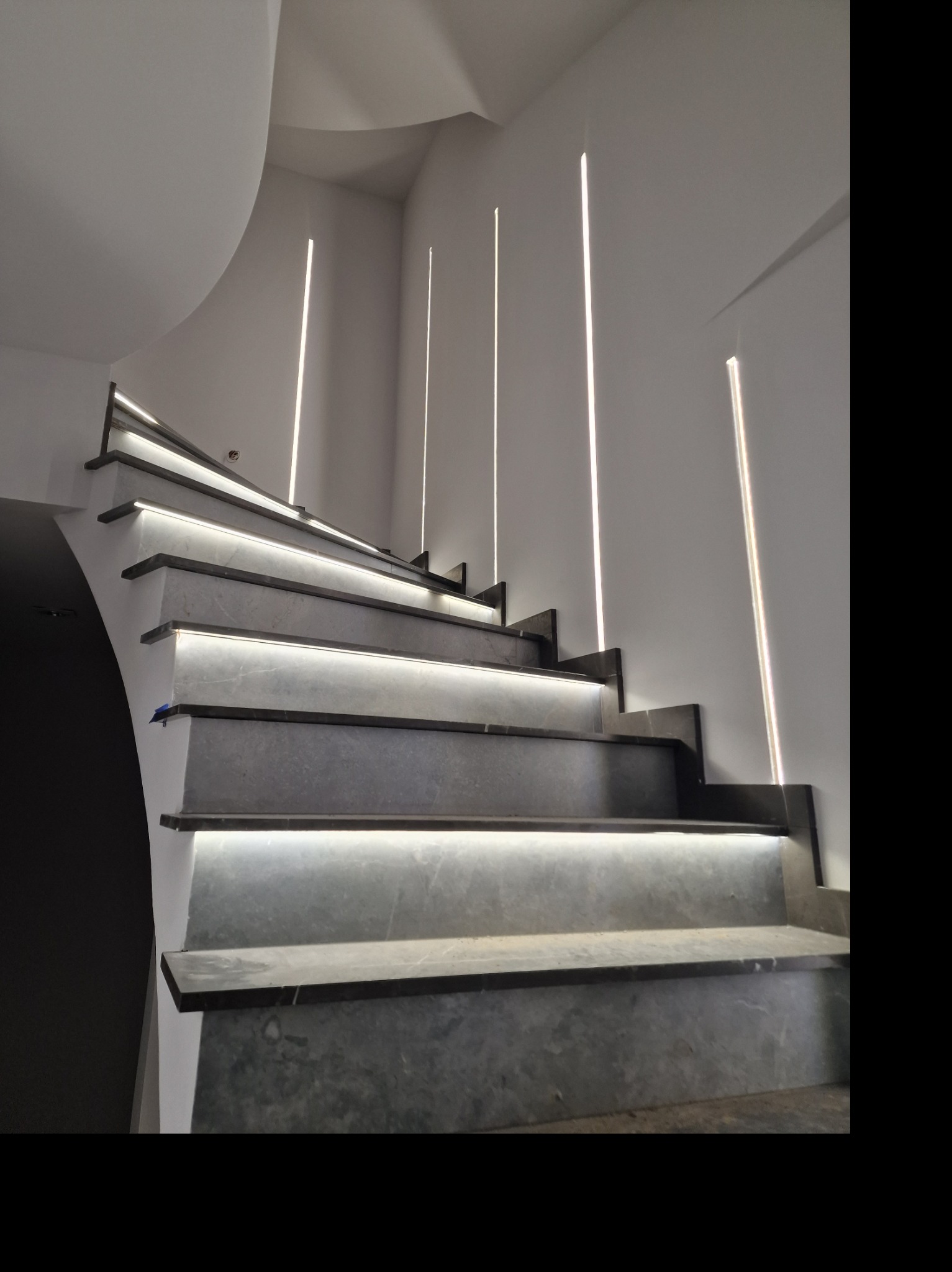Nowoczesne schody z podświetleniem LED na stopniach i pionowe linie świetlne na ścianie, tworzące unikalny efekt wizualny w minimalistycznym wnętrzu.