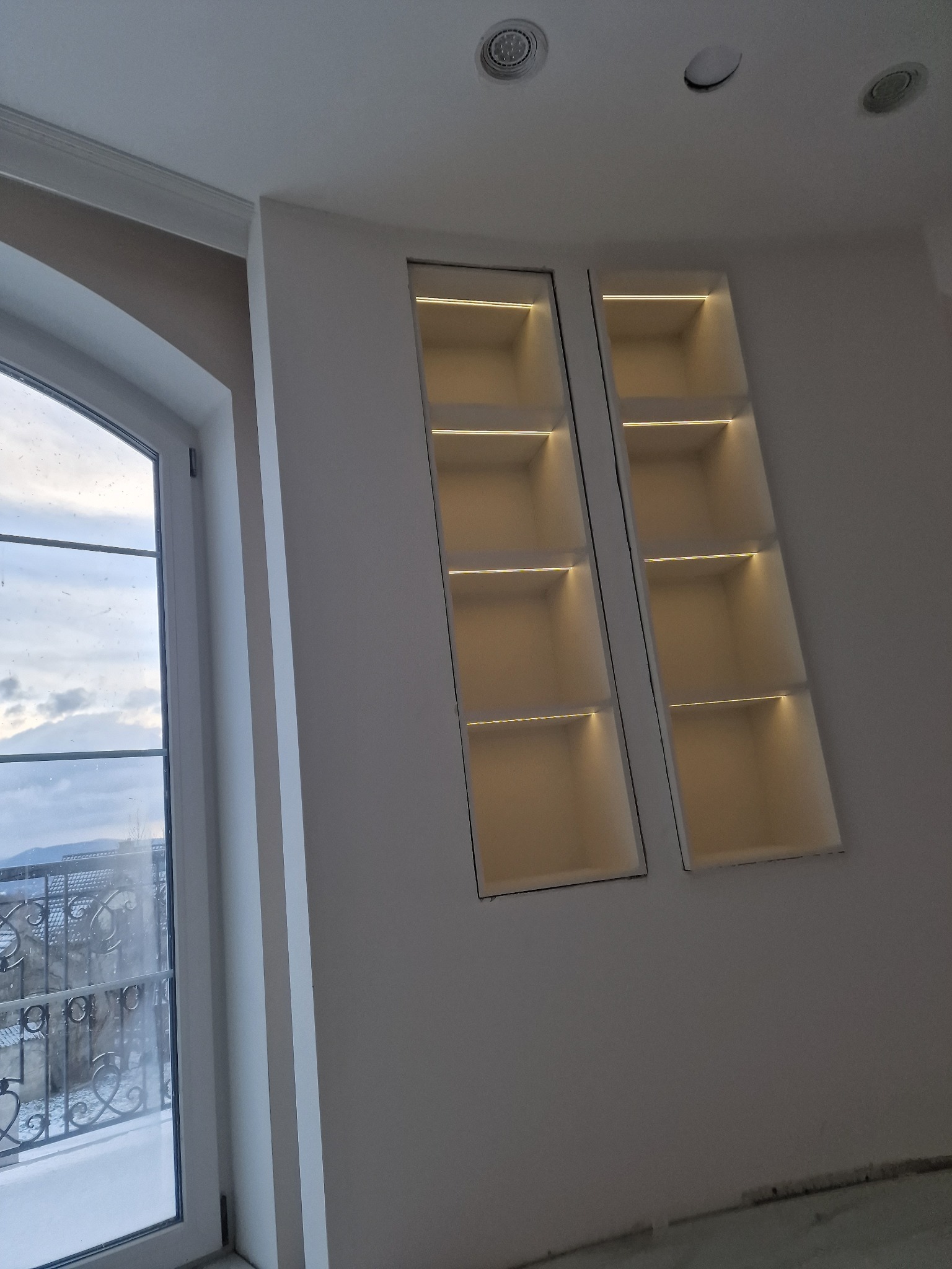 Dwa podświetlane, wnękowe regały na ścianie, z widokiem na zewnątrz przez okno. Oświetlenie LED w półkach tworzy ciepłą atmosferę.