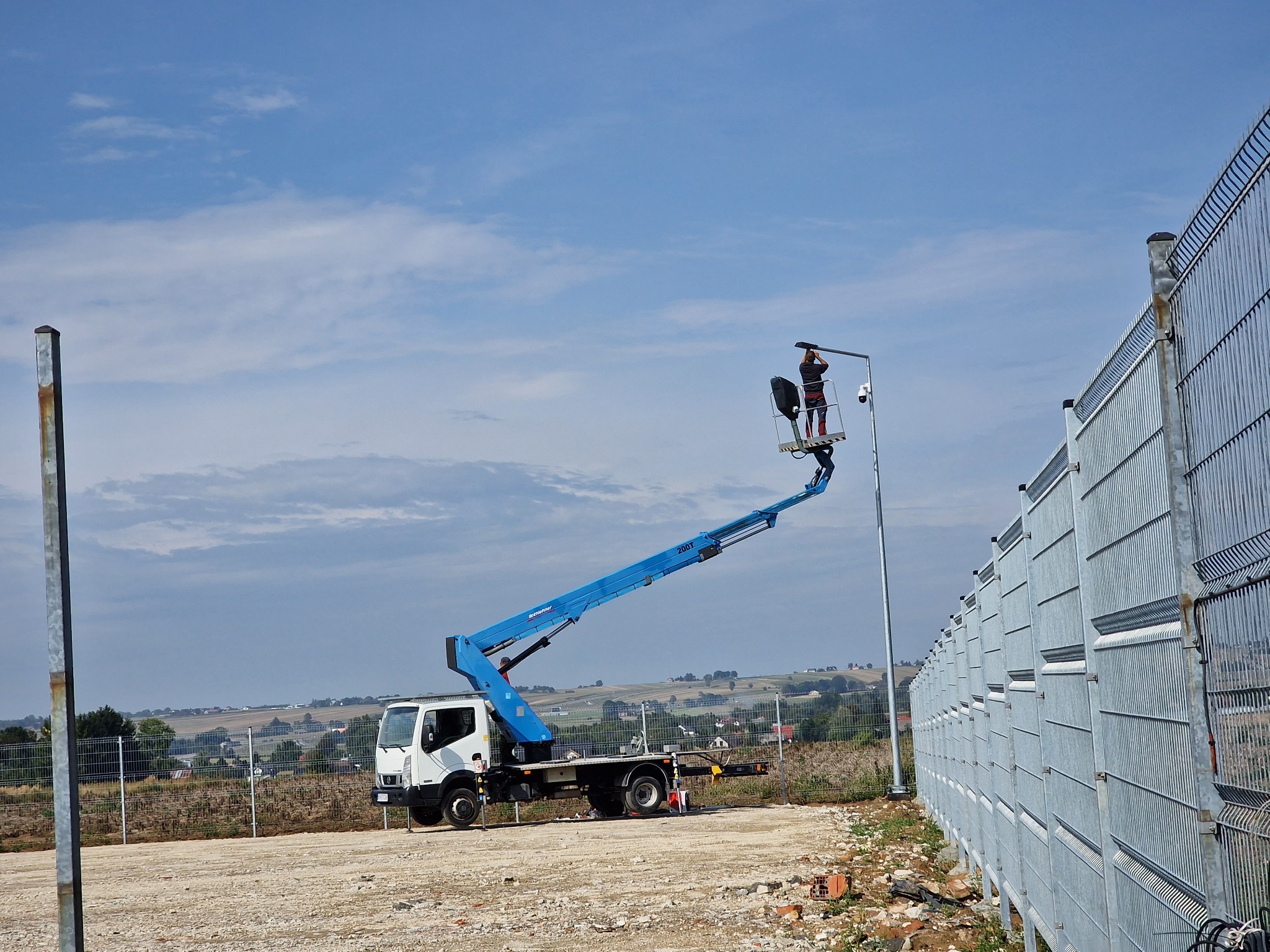 Pracownik na podnośniku montuje oświetlenie na słupie wzdłuż metalowego ogrodzenia. W tle krajobraz wiejski pod błękitnym niebem.