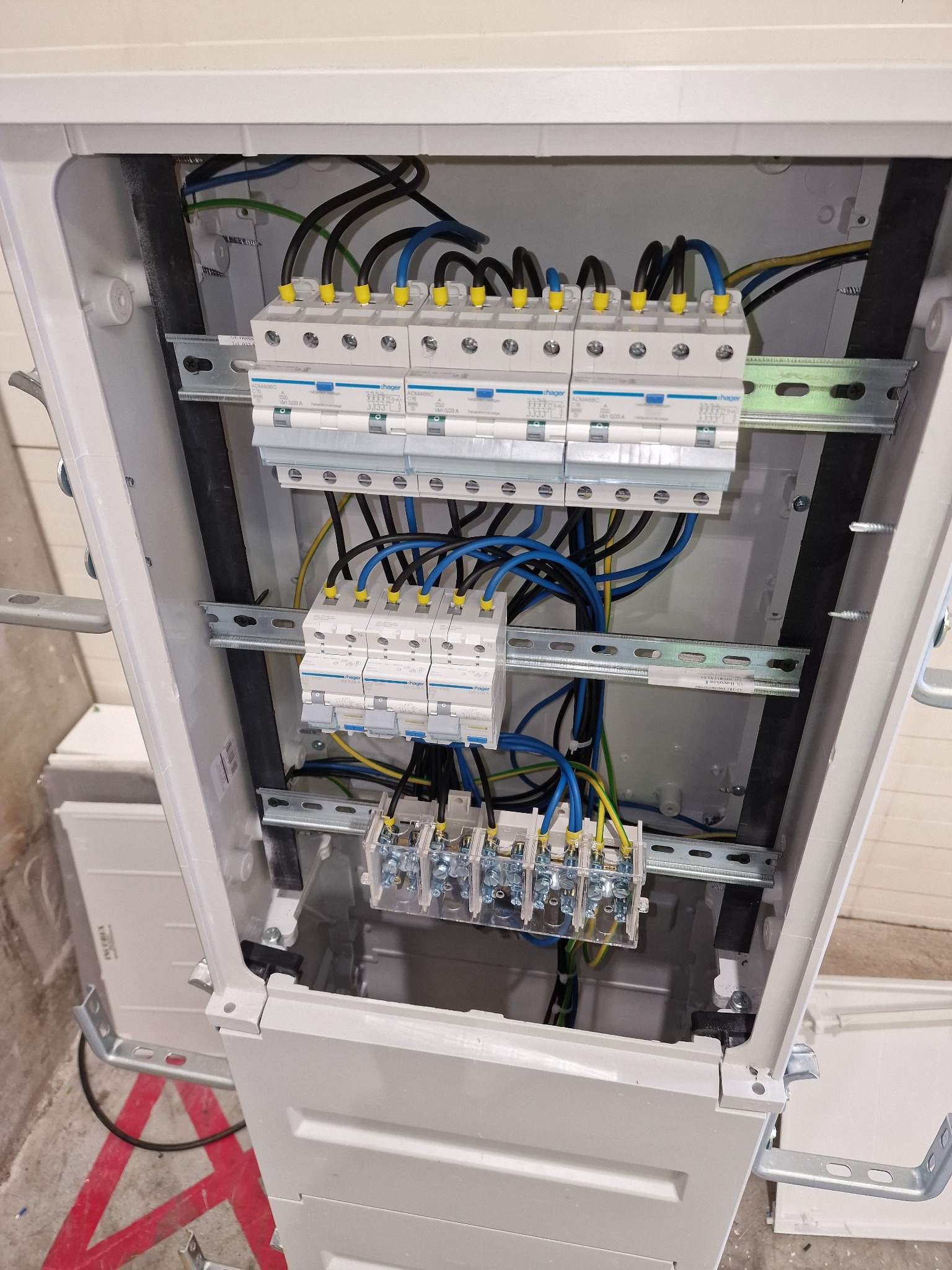 Wnętrze skrzynki elektrycznej z zamontowanymi bezpiecznikami i podłączonymi przewodami w kolorach: czarnym, niebieskim, żółto-zielonym. Widoczne szyny montażowe i elementy mocujące.