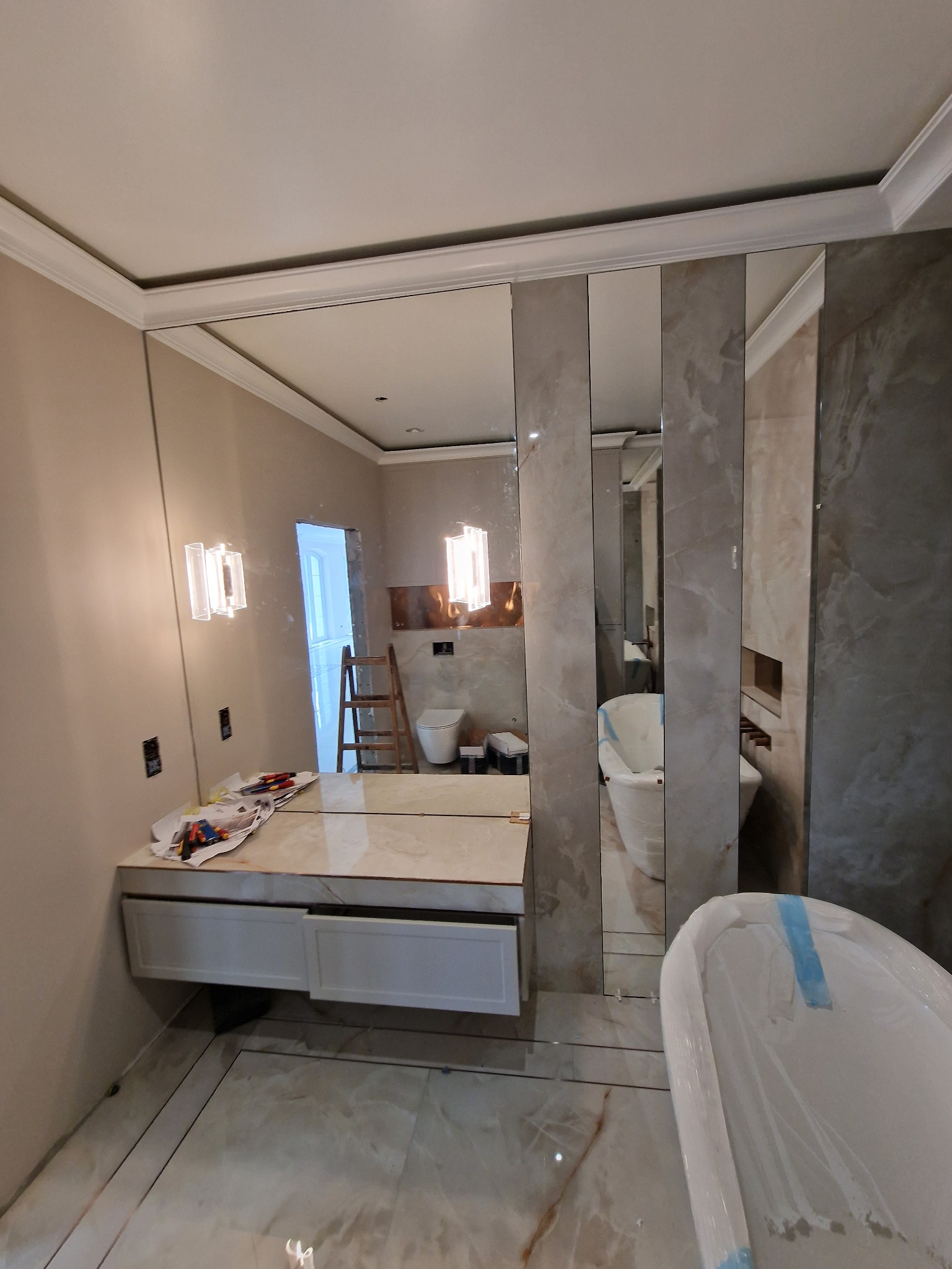 Nowoczesna łazienka w trakcie wykańczania: lustra w pionowych pasach, biała wanna zabezpieczona folią, oświetlenie ścienne i marmurowe płytki. Widoczne narzędzia.