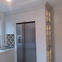 Biała zabudowa kuchenna z lodówką side-by-side i szafką z podświetlanymi, dekoracyjnymi wnękami. Widoczny fragment blatu i półka na wino.