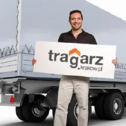 Uśmiechnięty mężczyzna prezentuje baner z logo 'tragarz.krakow.pl' na tle srebrnej naczepy ciężarówki z plandeką. Widoczne tylne światła i koła.