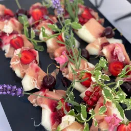 Elegancka kompozycja przekąsek na czarnym talerzu: melon owinięty szynką parmeńską, udekorowany truskawkami, czereśniami, czerwoną porzeczką i świeżymi ziołami, tworząca apetyczną prezentację.