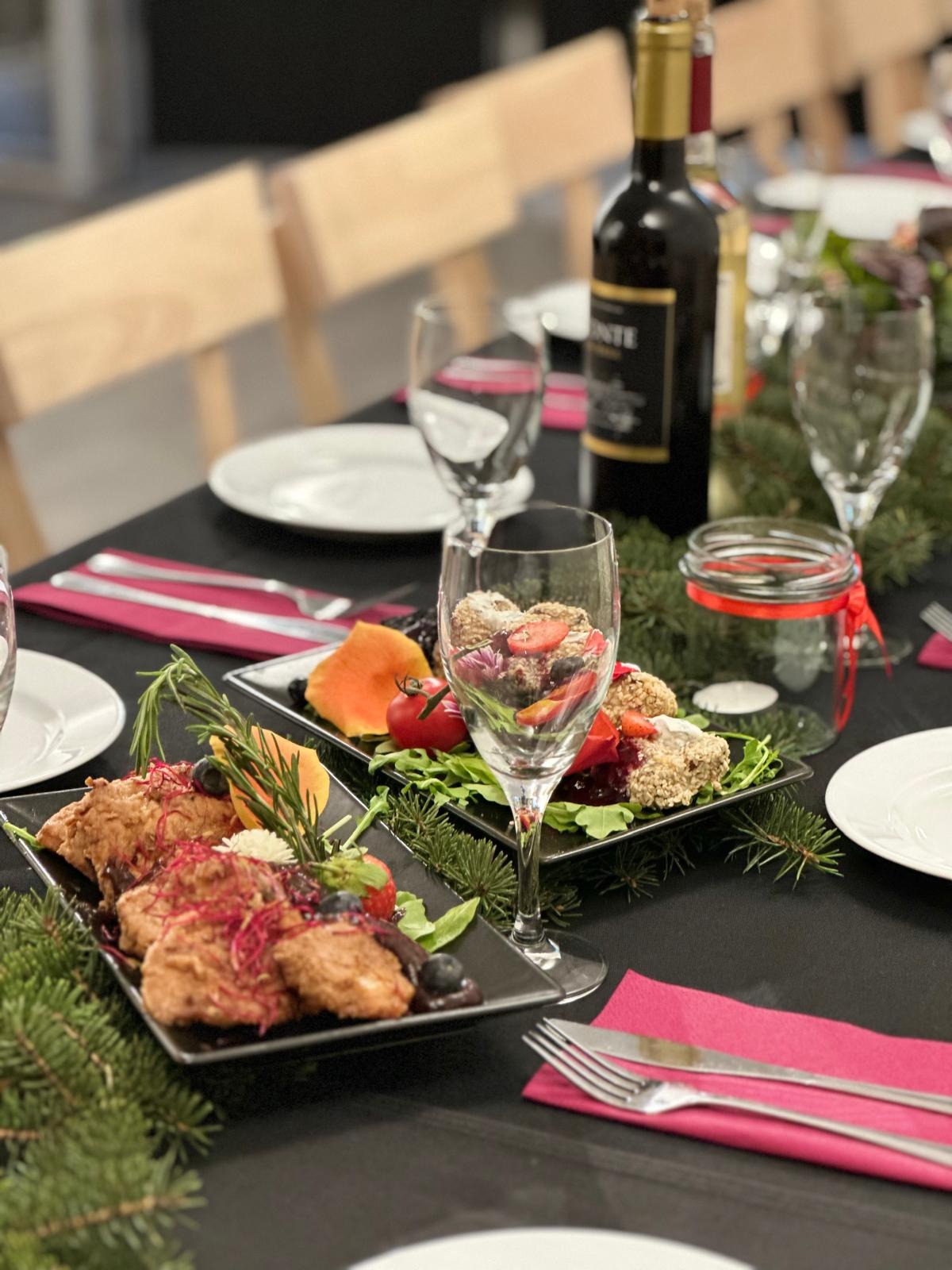 Uroczysty stół z czarnym obrusem, zastawiony talerzami z przekąskami, owocami i ziołami, kieliszkiem z deserem, butelką wina i świątecznymi gałązkami świerku.