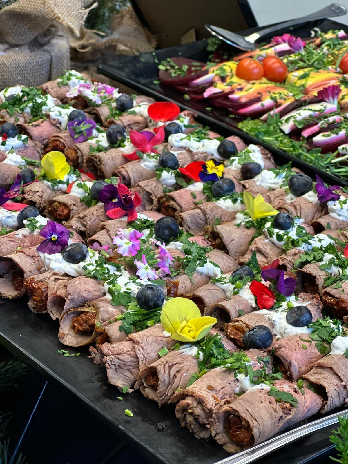 Roladki z rostbefu z kremowym serem, borówkami i jadalnymi kwiatami na czarnym prostokątnym talerzu, obok sałatka z buraków i pomidorów.