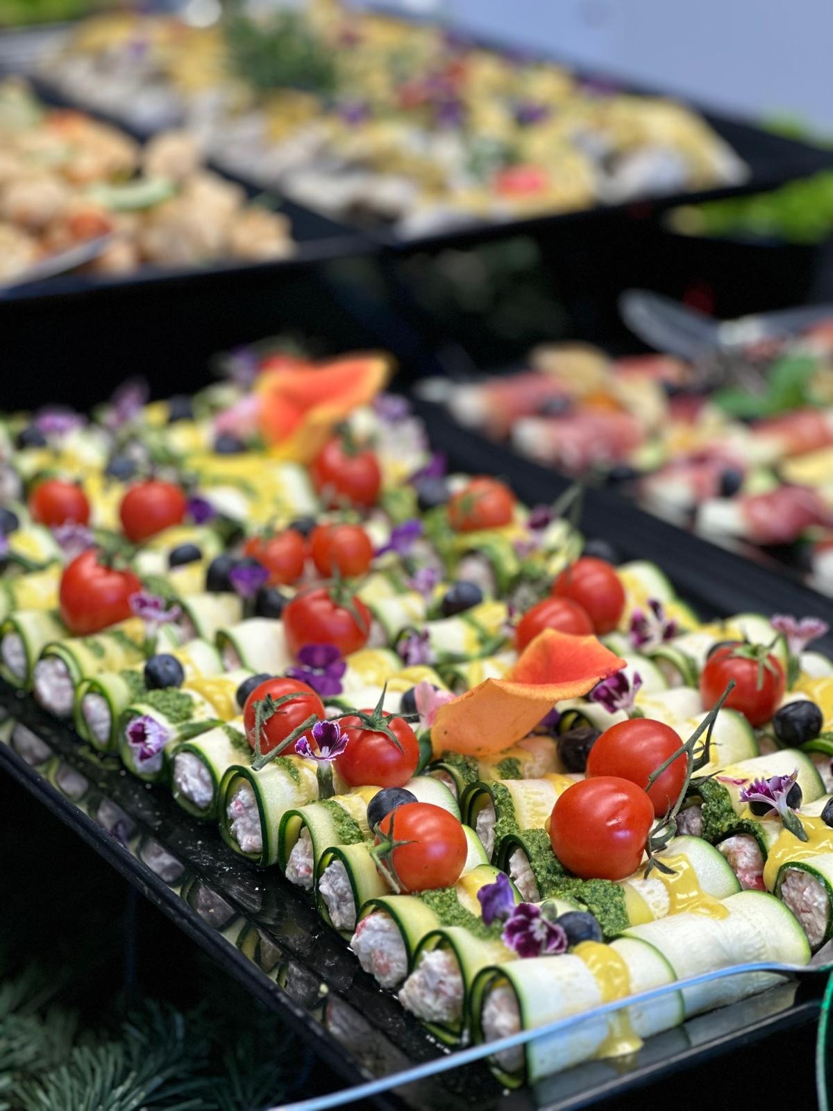 Elegancko podane roladki z cukinii z kremowym nadzieniem, udekorowane pesto, pomidorkami cherry, borówkami i jadalnymi kwiatami na czarnym talerzu, część bufetu w tle.