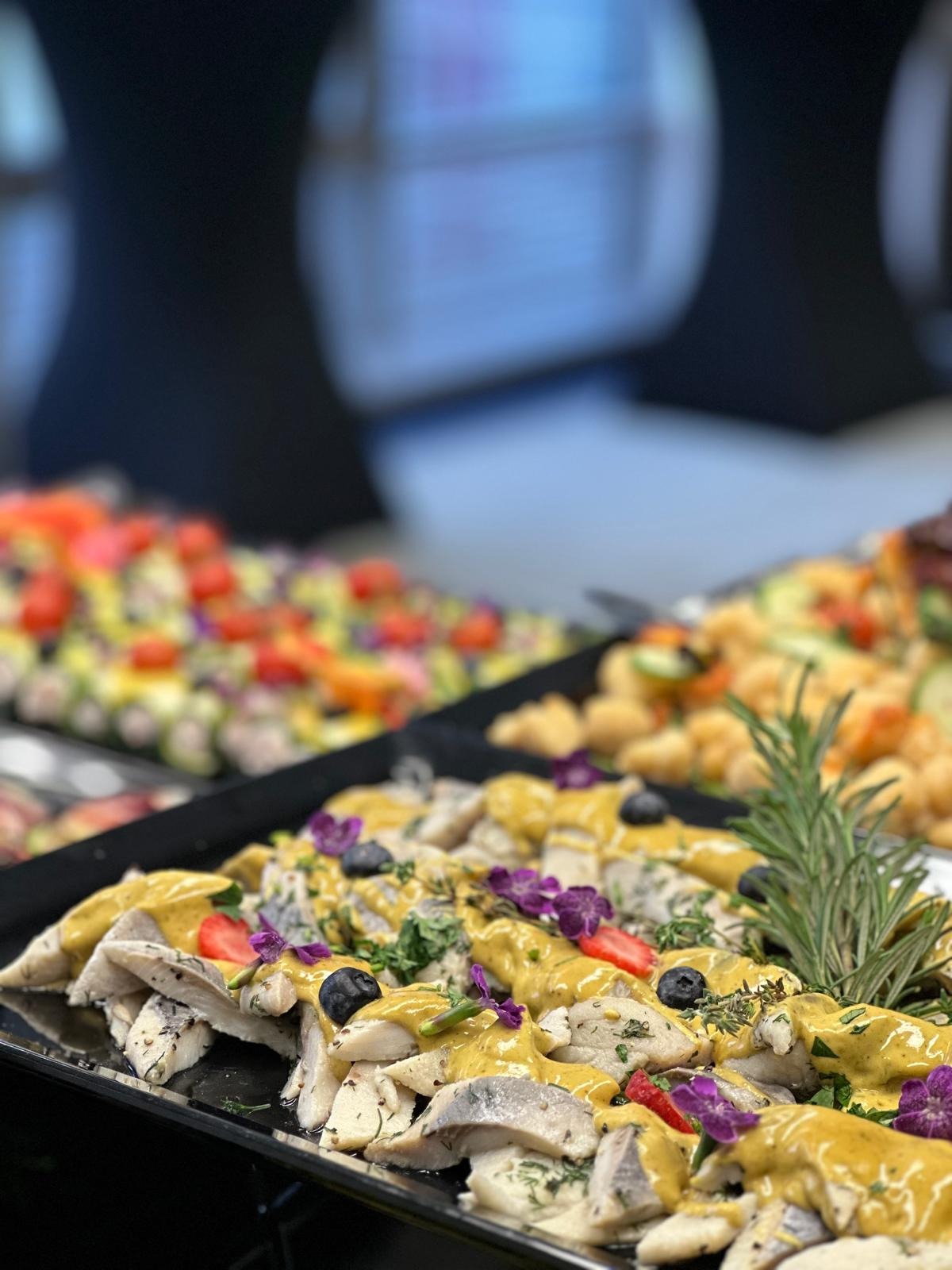 Elegancko udekorowany półmisek z śledziem w sosie musztardowym, ozdobiony borówkami, truskawkami, ziołami i fioletowymi kwiatami, na tle innych potraw.