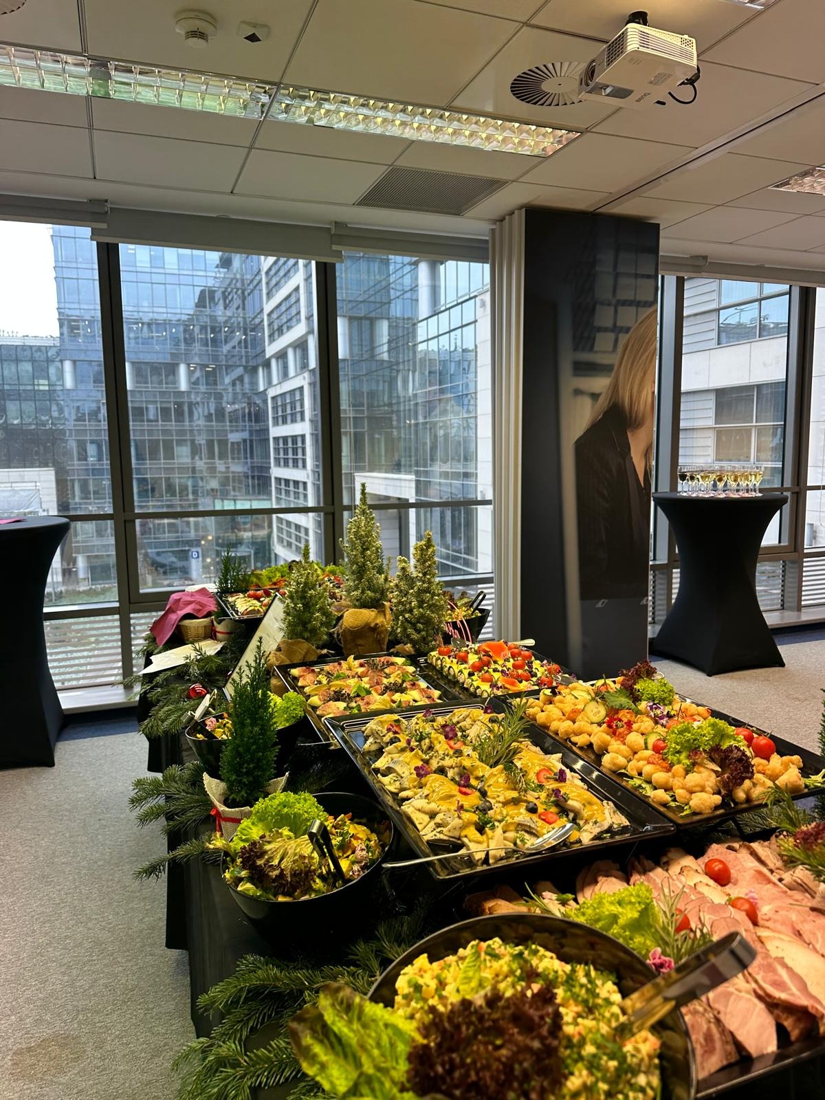 Bogato zastawiony stół cateringowy z różnorodnymi daniami, dekoracjami świątecznymi w tle biurowiec z dużymi oknami i projektor na suficie.