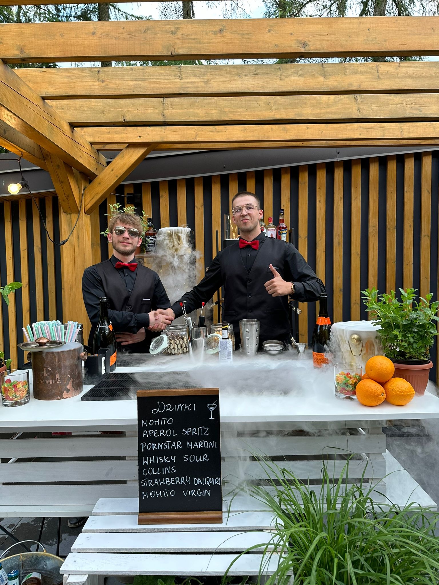 Dwóch barmanów w czarnych koszulach i czerwonych muchach za białym barem z dymem, oferujących drinki z menu na tablicy, w tle drewniana konstrukcja.