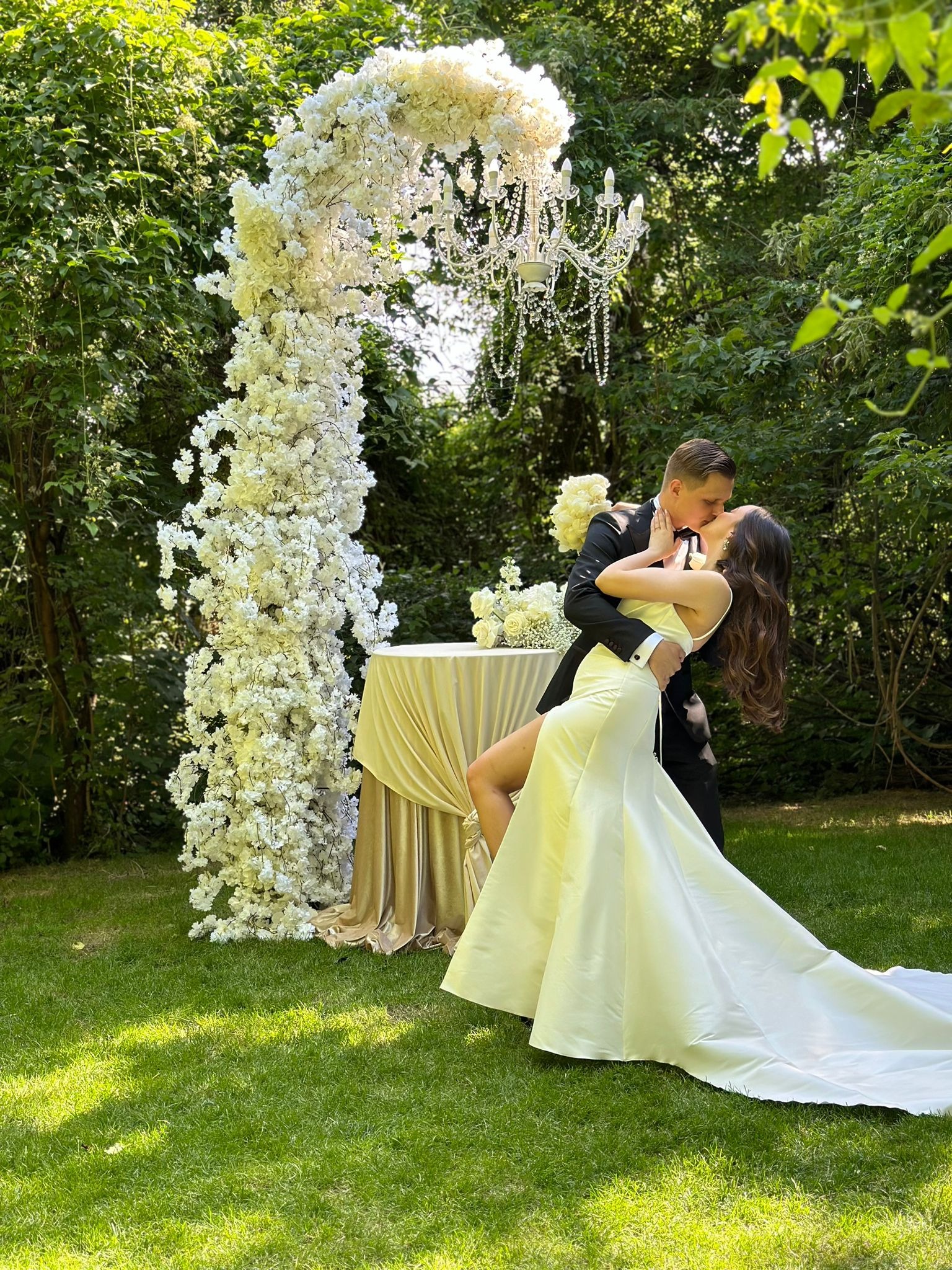 Para młoda w Warszawie całuje się pod kwiatową bramą z kryształowym żyrandolem w tle, na trawniku. Dekoracja stołu z kwiatami w oddali.