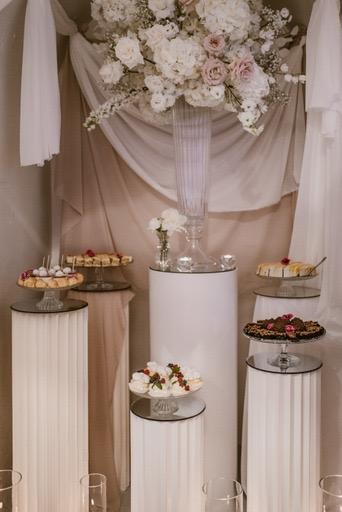 Elegancki bufet z deserami na białych kolumnach, udekorowany kwiatami w odcieniach bieli i różu, z beżowym drapowanym tłem.
