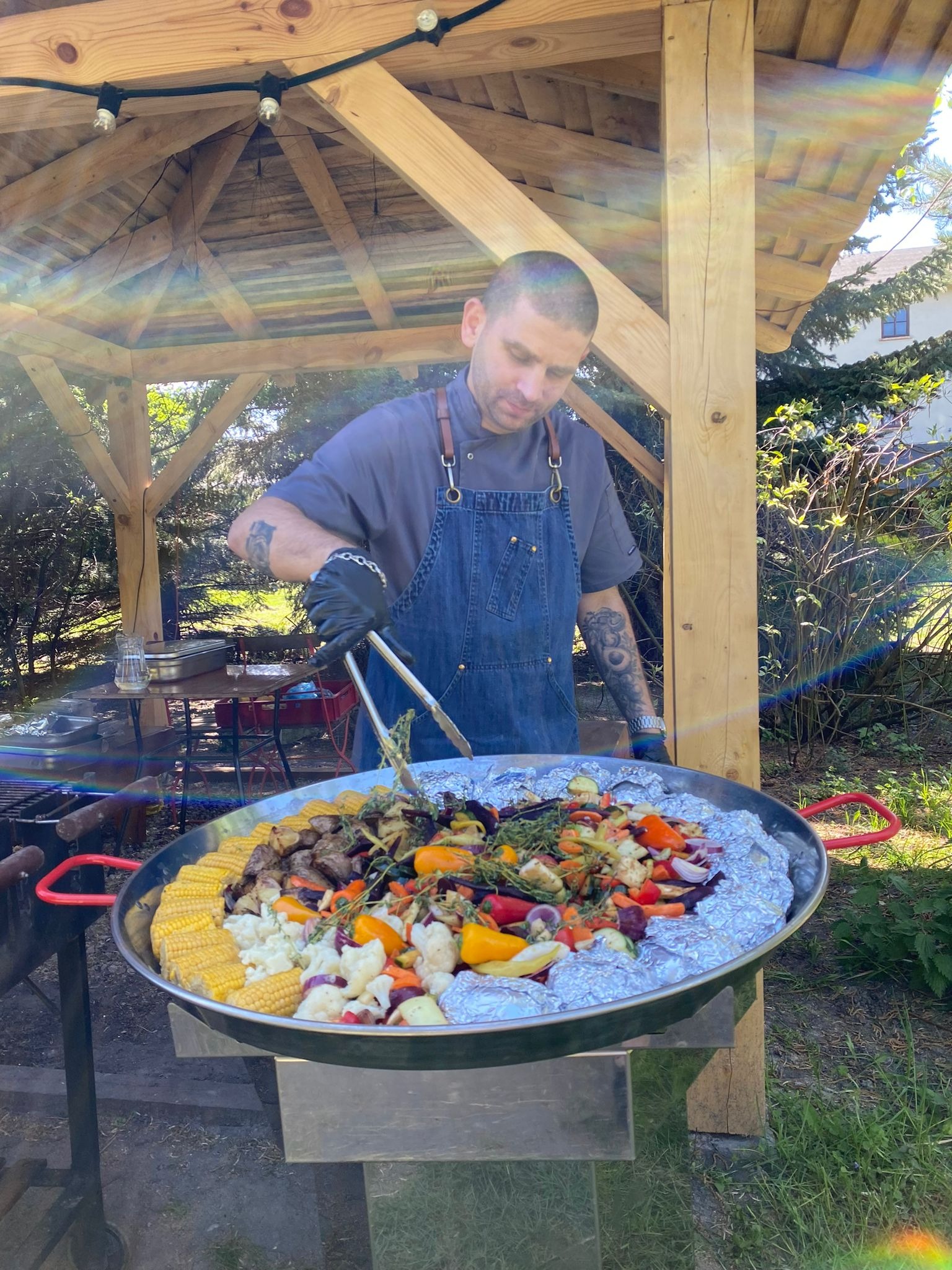 Kucharz w denimowym fartuchu i czarnych rękawicach, przygotowuje paellę z kukurydzą, warzywami i mięsem na świeżym powietrzu w dużej patelni, pod drewnianą altaną.