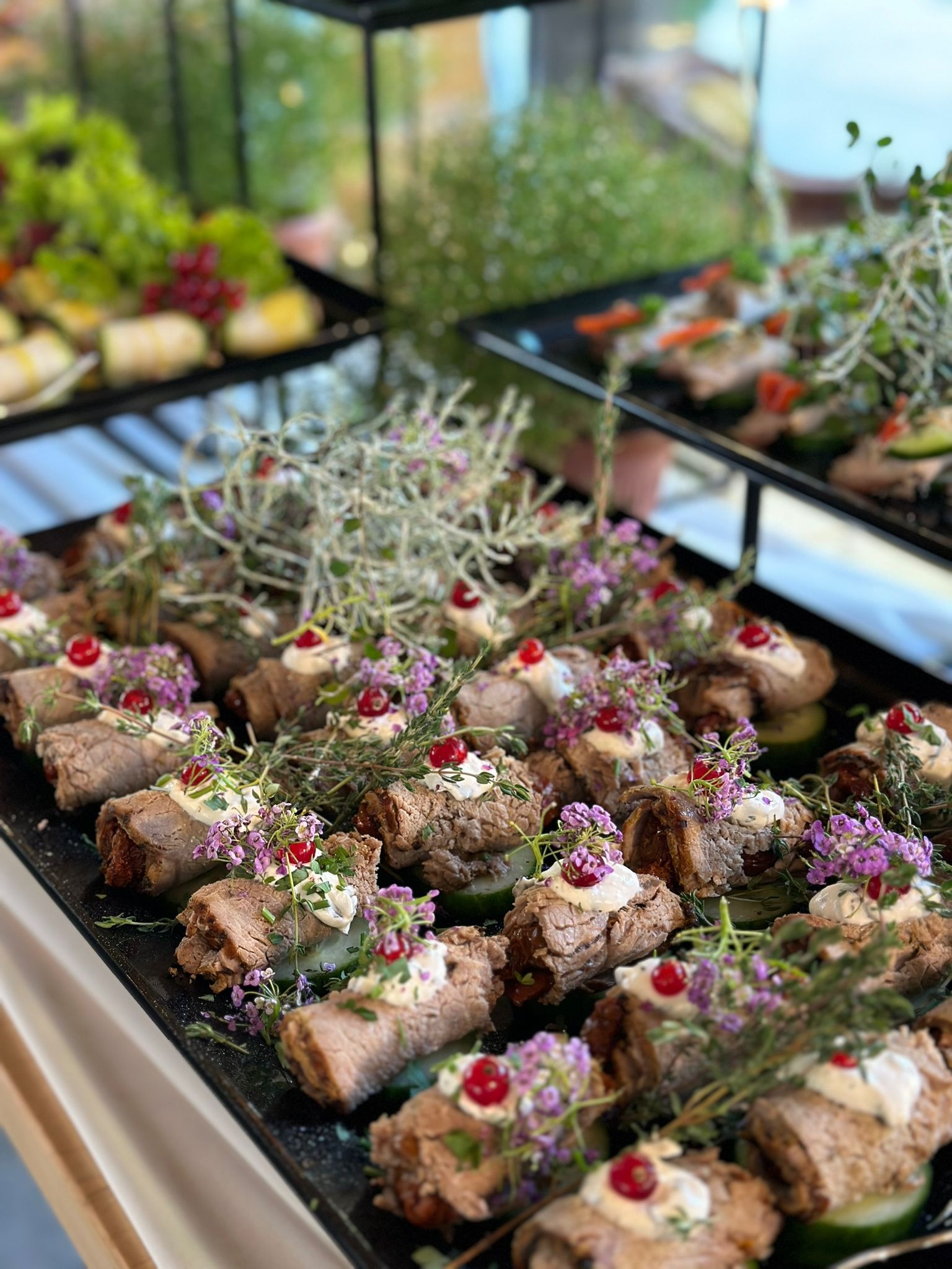 Elegancko podane roladki z mięsa z kremowym sosem, czerwoną porzeczką i gałązką ziół na plastrach ogórka, ułożone na czarnej tacy cateringowej.