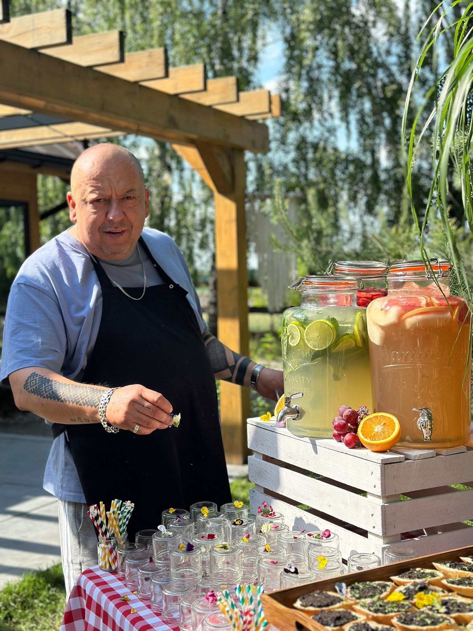 Mężczyzna w czarnym fartuchu dekoruje kwiatami szklanki obok trzech dużych słojów z lemoniadą owocową na rustykalnym, białym podeście, widoczne również kanapeczki na drewnianej tacy.