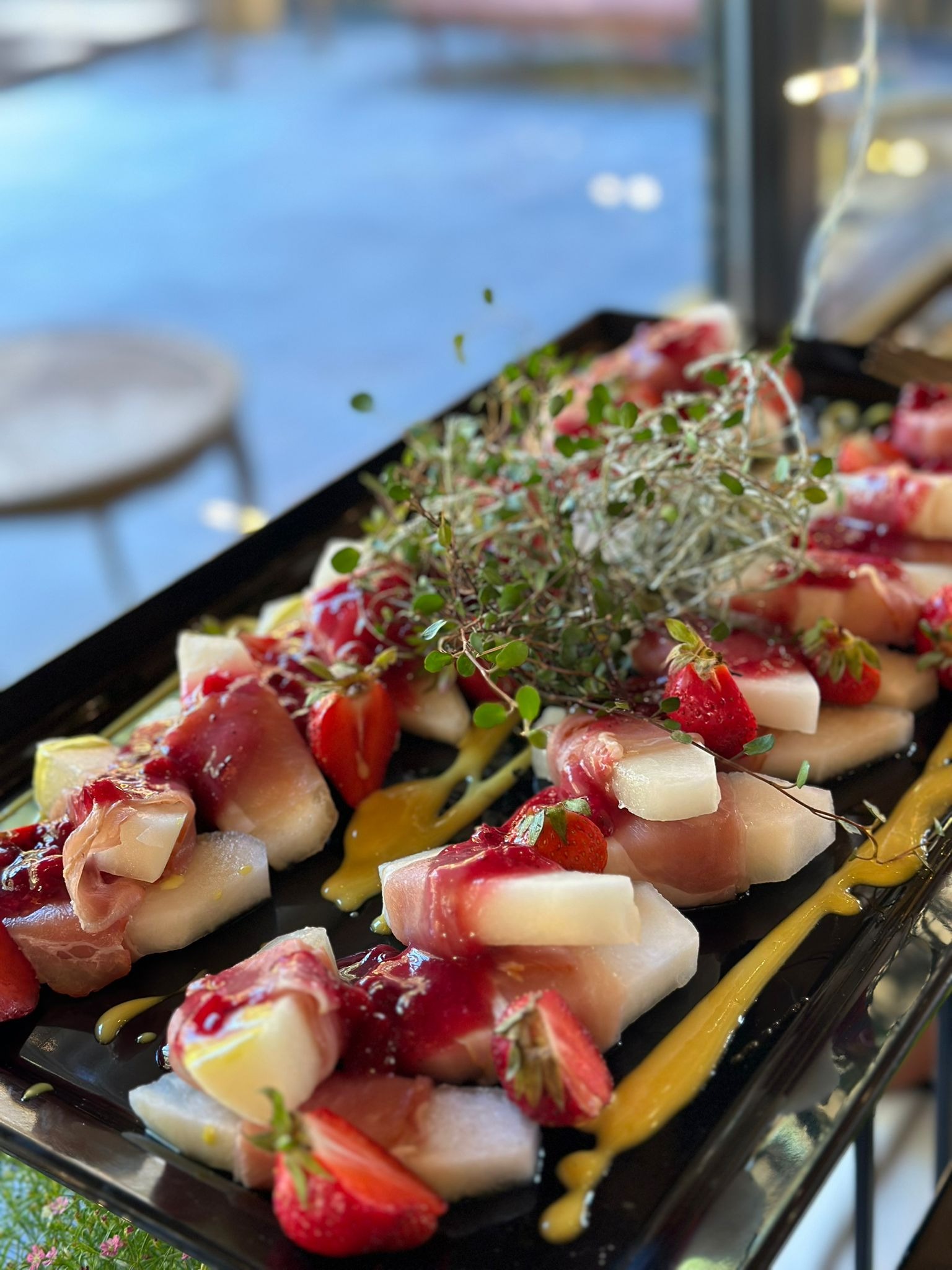 Elegancka przekąska na czarnym talerzu: kawałki melona owinięte szynką parmeńską, udekorowane sosem malinowym, świeżymi truskawkami i gałązką ziół.