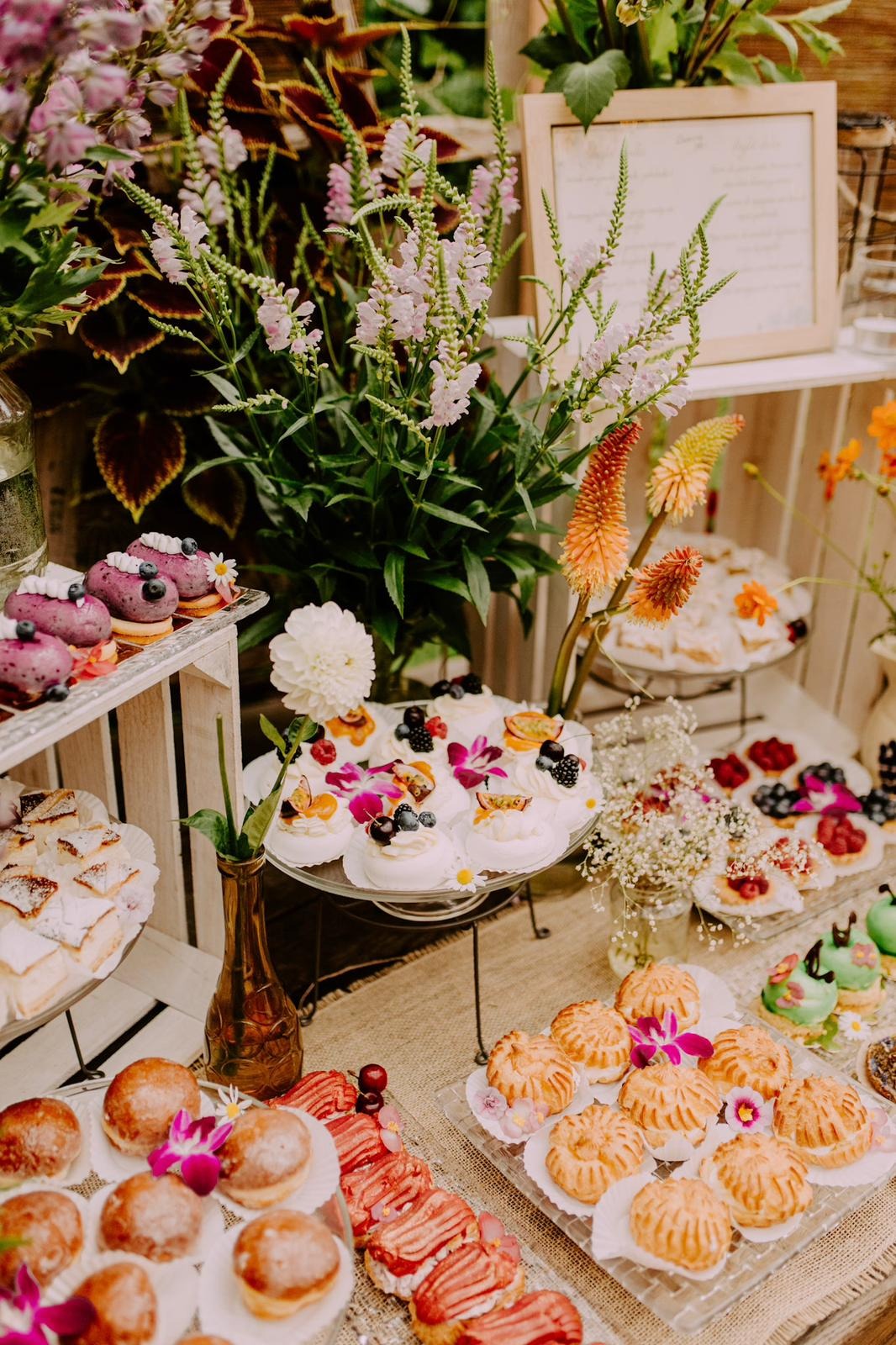 Bogato zastawiony stół z różnorodnymi deserami, w tym mini bezy z owocami, pączki, eklerki, ciasta i babeczki, udekorowany kwiatami i zielenią, tworzący elegancką kompozycję.