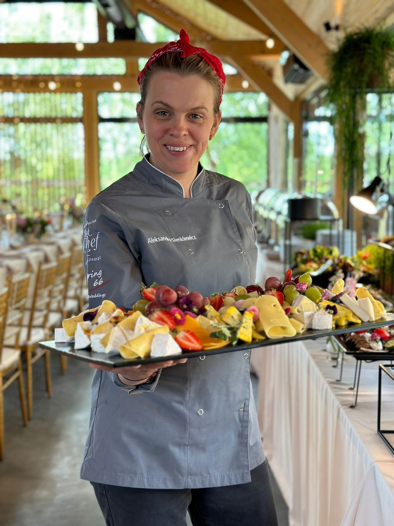 Uśmiechnięta szefowa kuchni w szarym uniformie prezentuje tacę z różnorodnymi serami i owocami, w tle sala bankietowa z drewnianą konstrukcją dachu.