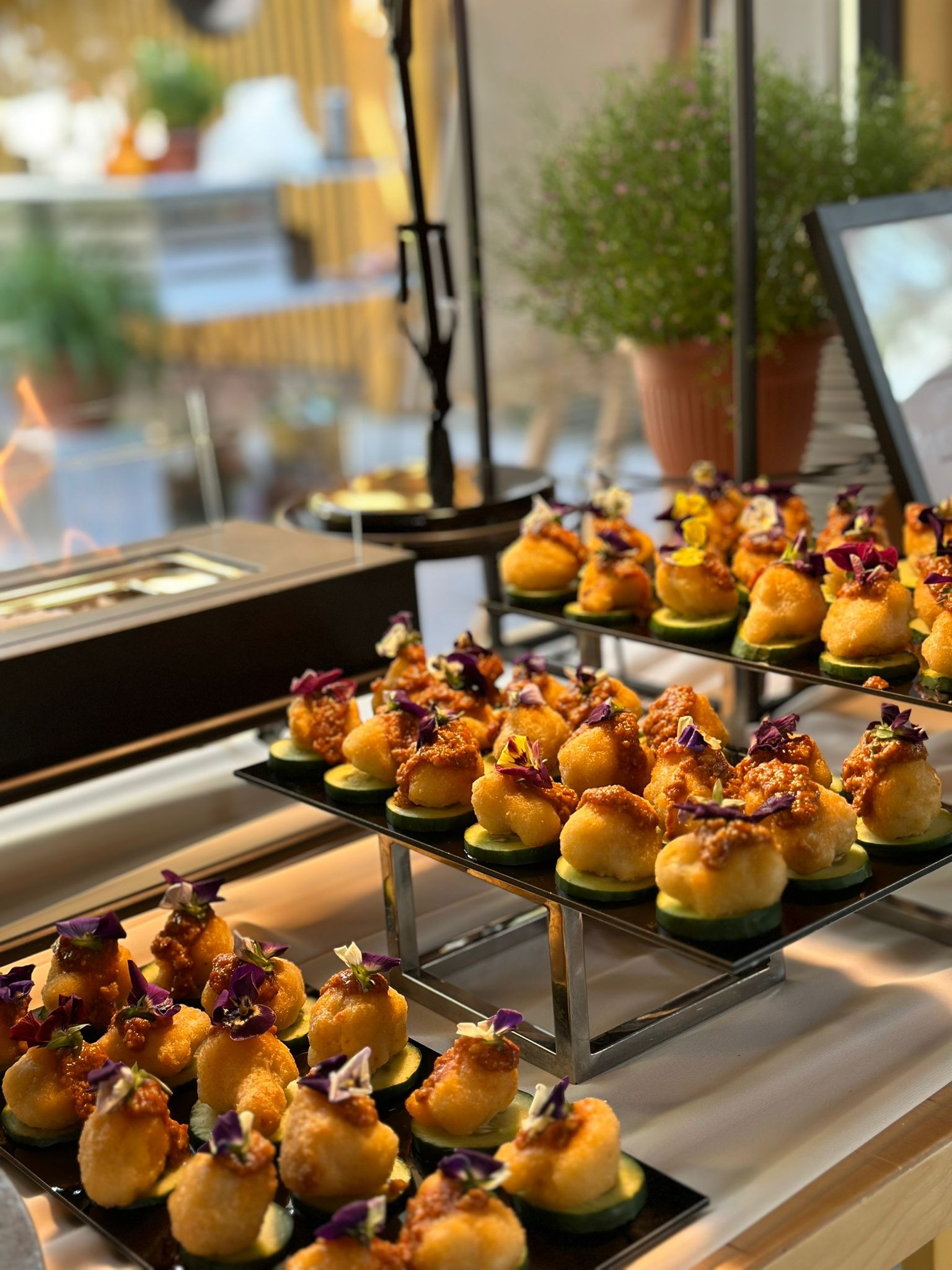 Elegancko zaaranżowane, złociste przekąski na plastrach ogórka, udekorowane jadalnymi kwiatami, ułożone na czarnych, lustrzanych tacach, stanowiące wyrafinowany element cateringu.