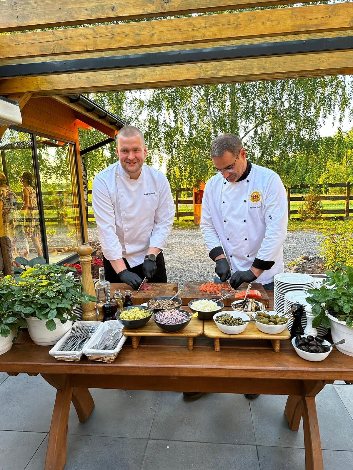 Dwóch kucharzy w białych uniformach przygotowuje tatara na drewnianym stole z dodatkami, widoczne naczynia z kaparami, oliwkami i posiekanymi warzywami, w tle zieleń ogrodu.