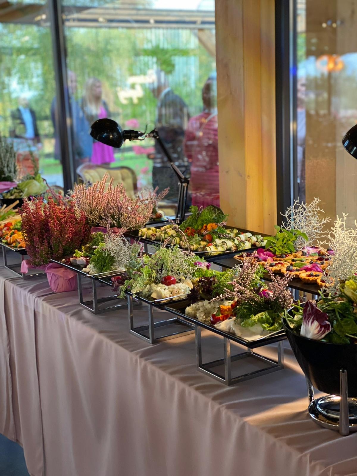 Elegancko zaaranżowany bufet z różnorodnymi sałatkami i przekąskami, udekorowany wrzosami i oświetlony lampkami, widoczni goście w tle.