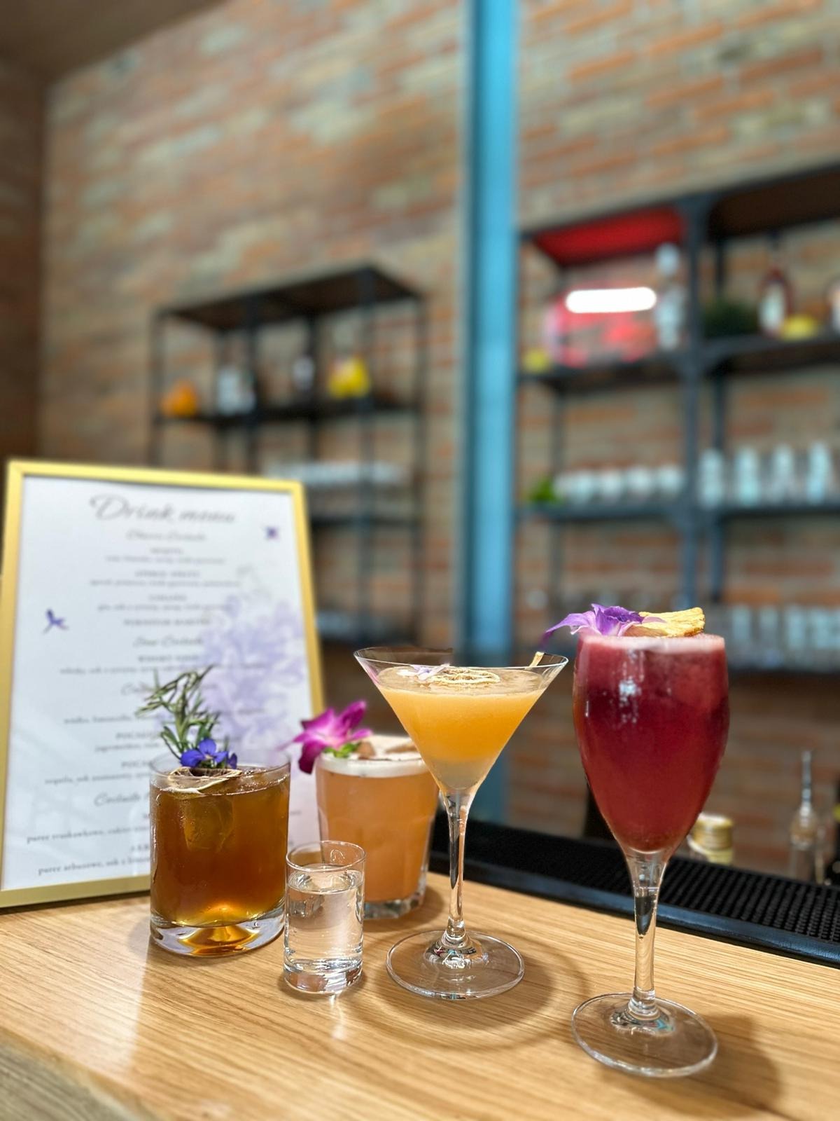 Cztery kolorowe koktajle na drewnianym barze, udekorowane kwiatami i ziołami, z menu drinków w złotej ramce w tle, na tle ceglanej ściany i metalowych półek.
