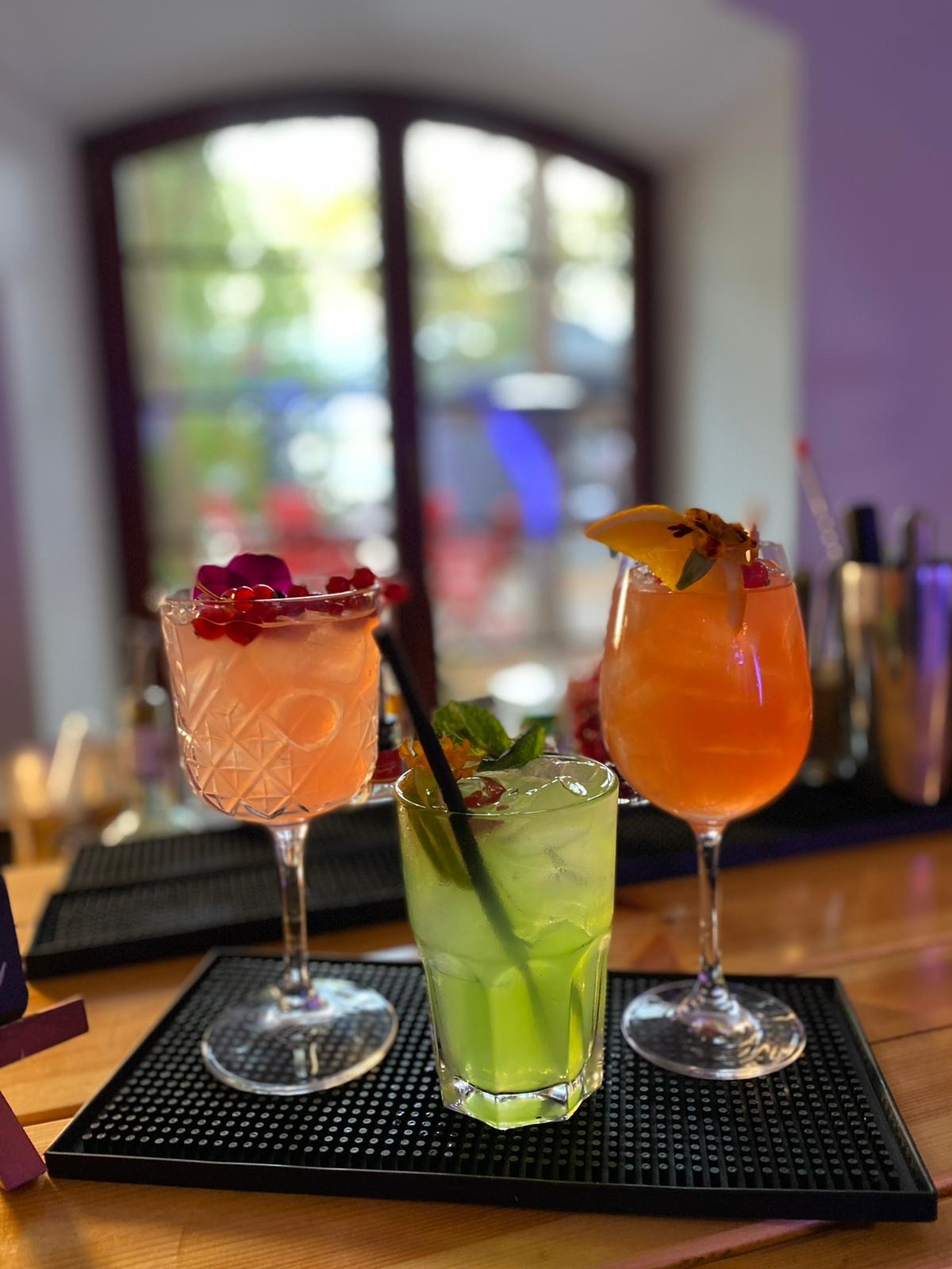 Trzy kolorowe koktajle na barze, ozdobione owocami i kwiatami, podane na czarnej podkładce z widocznym oknem w tle.