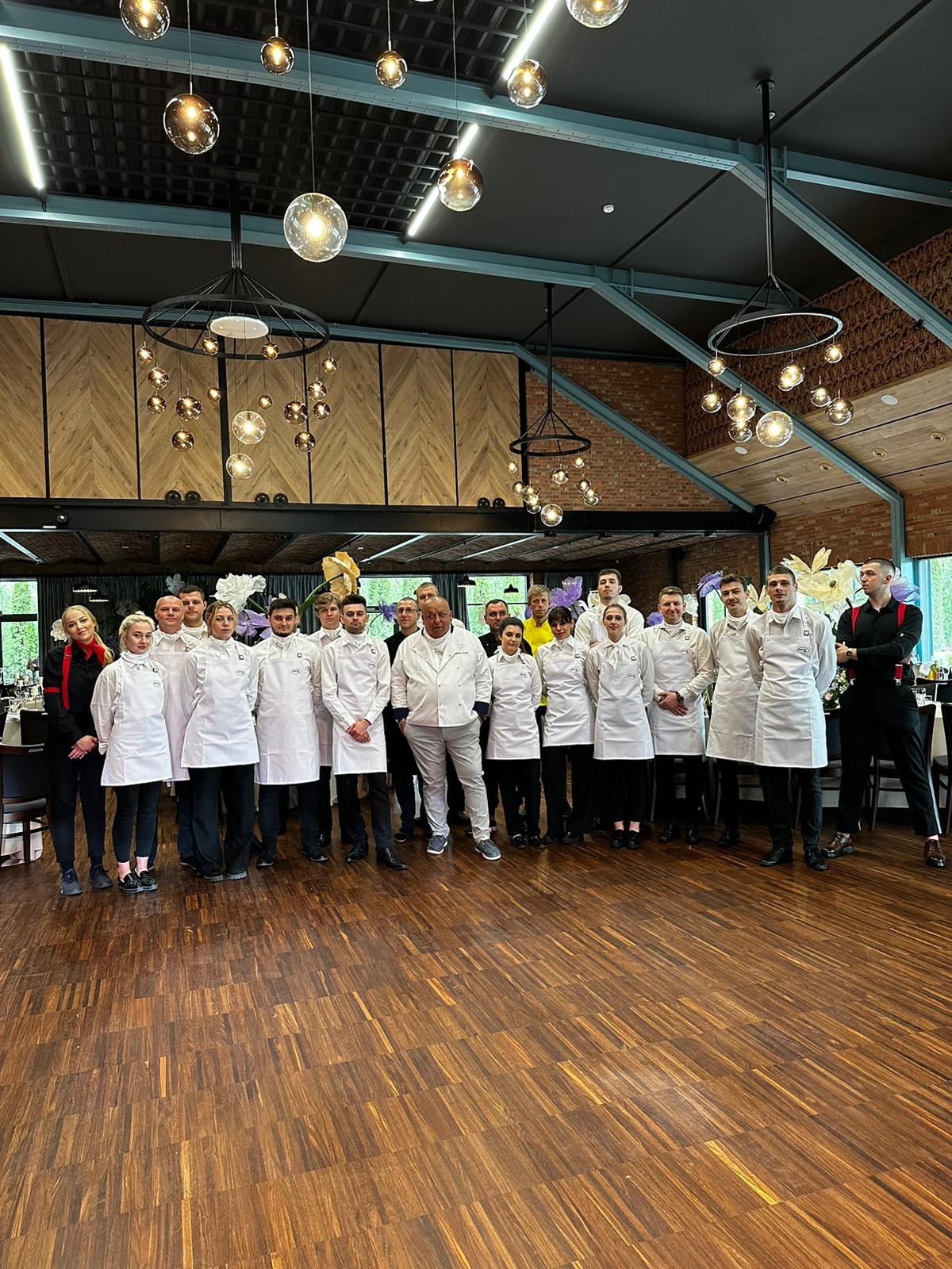 Zespół kucharzy i kelnerów w białych uniformach i fartuchach, stojący na parkiecie w przestronnym, eleganckim wnętrzu restauracji z dekoracjami kwiatowymi.